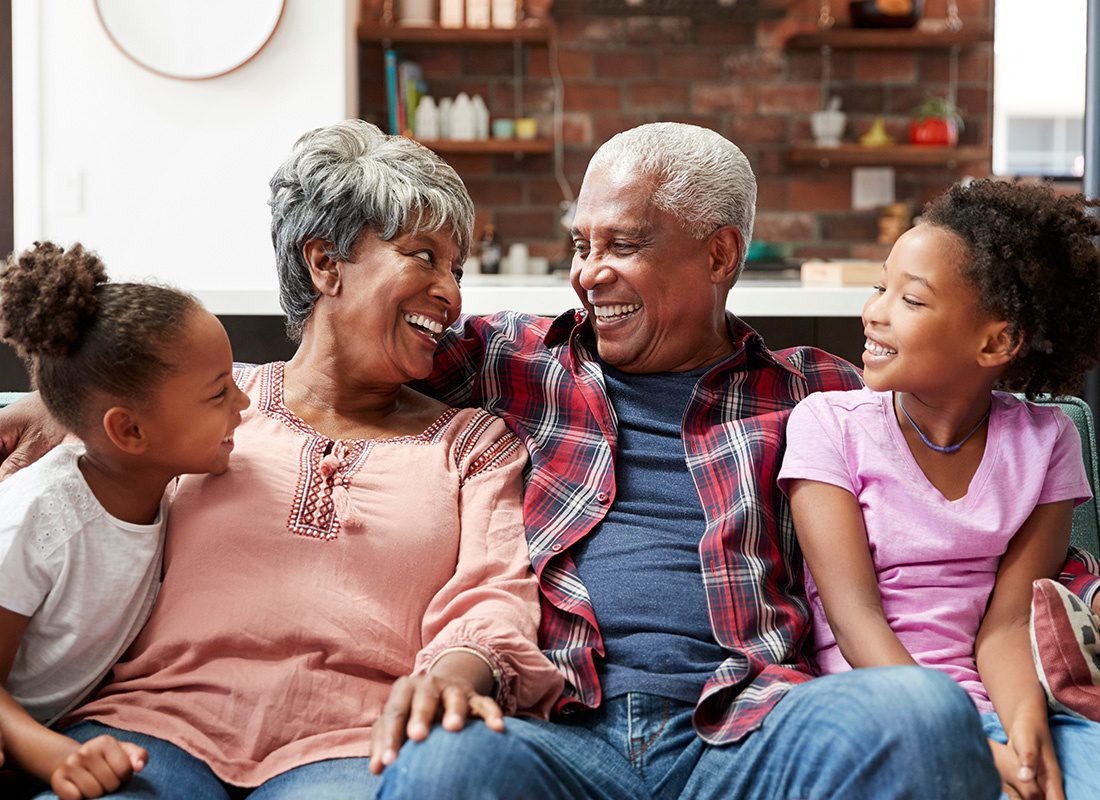 About Our Agency - Grand Parents and Their Two Young Grand Daughters Sit and Laugh Together at Home